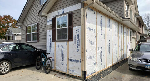 A home exterior with new insulation panels stacked by the wall and a bicycle leaning beside a parked car in the driveway.