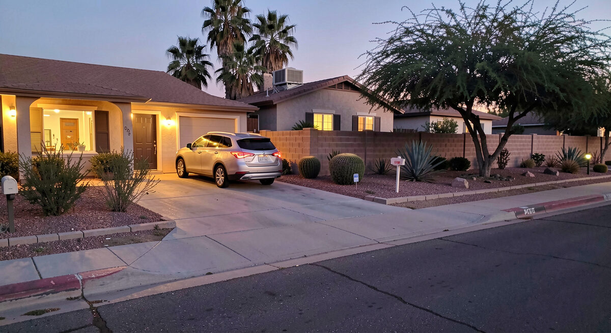 A family home at dusk with warm lights on and a parked car in the driveway, desert shrubs along the wall.